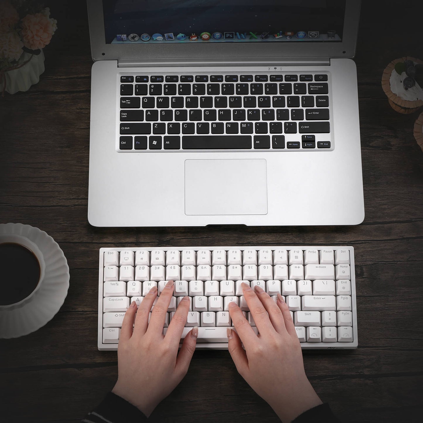 A user typing on a Royal Kludge RK84 compact gaming keyboard placed on a wooden table, alongside a laptop, a cup of coffee, and a decorative plate. The keyboard features a sleek white design and is positioned for easy access.