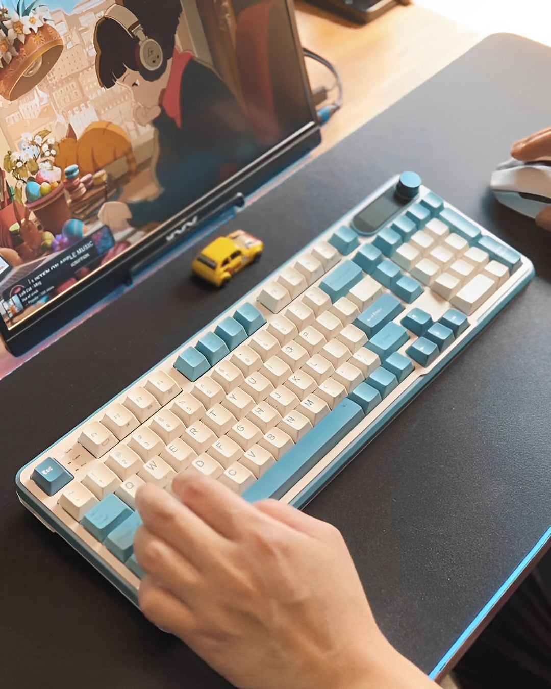 Hand using a Royal Kludge S98 keyboard with a sleek design and pastel blue accents, placed on a desk with a computer monitor and gaming elements